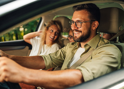 Un homme barbu portant des lunettes conduit une voiture, une femme souriante à ses côtés et un enfant assis à l’arrière-plan, en plein départ en vacances. Un homme barbu portant des lunettes conduit une voiture, une femme souriante à ses côtés et un enfant assis à l’arrière-plan, en plein départ en vacances.